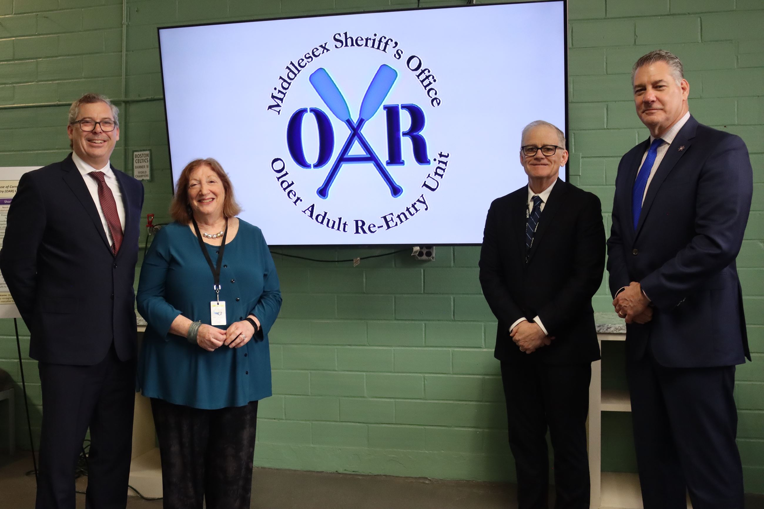 Four people stand in front of a TV with a logo for the Older Adult Re-Entry Unit.